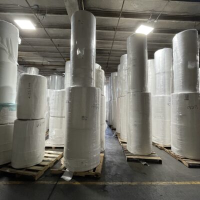 White nonwoven parent rolls stacked in a warehouse setting with a gray floor and ceiling lights.